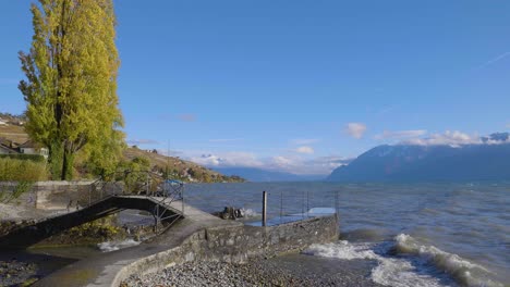 Waves-crashing-on-small-port-entrance-with-small-pathway-over-bridge-Shores-of-Lake-Léman,-the-Alps-in-the-background