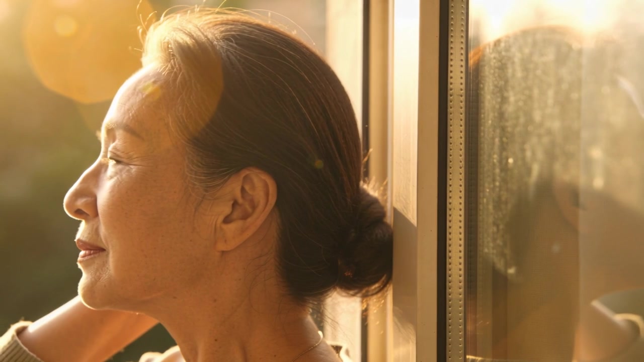 ガラスのそばで明るいトップスを着たアジア人女性が寄りかかり、太陽が顔を照らす中、お団子ヘアを直している。反射とレンズフレアが見える。