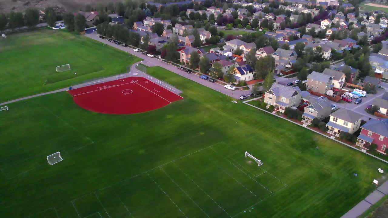 A zoom out shot of a baseball field in the suburbs. next to train tracks and an empty field .