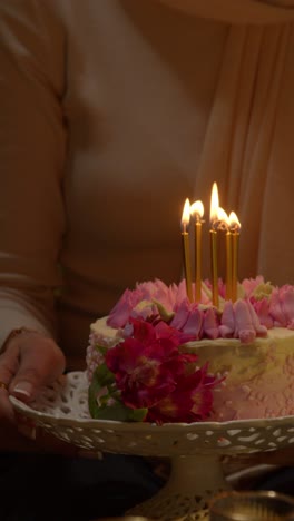 video vertical de cerca de una mujer con hijab sosteniendo un pastel de cumpleaños decorado con velas encendidas en una fiesta en casa