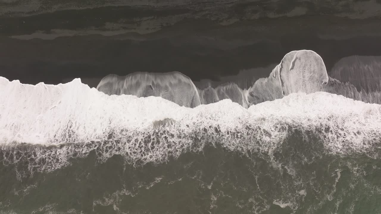 Aerial shot of waves crashing against the black sand beach of Reynisfjara, Iceland, highlighting the powerful contrast between the white surf and dark volcanic sand.