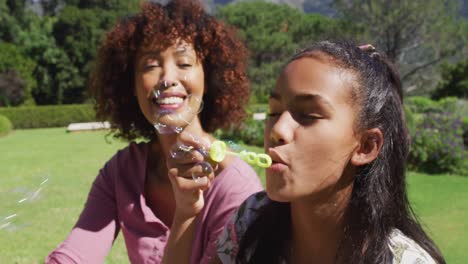madre y hija afroamericanas felices sentadas al aire libre en una manta, soplando burbujas