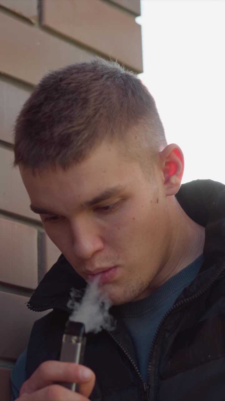 Close up of man using electronic cigarette outdoors near brick wall. subject holds and activates vaping device with consistent background lighting and soft focus on facial features and hand