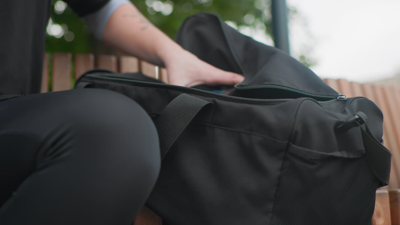 white person opens black duffel bag outdoors revealing contents, focused hands unzip wide, benchside scene with soft overcast light frames show athlete packing gym kit, photographer stowing camera