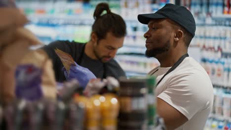 Close-up-shot-from-the-side-of-a-man-with-Black-skin-with-a-beard-in-a-white-T-shirt-in-a-black-apron-lays-out-and-sorts-a-counter-with-plastic-bottles-and-drinks-in-a-large-grocery-supermarket