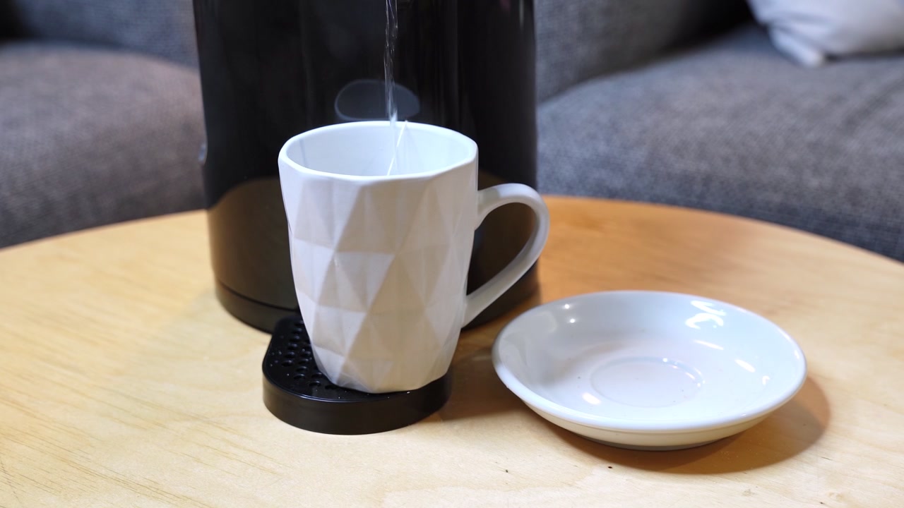 En kvinne med gullsmykker plasserer et hvitt geometrisk krus i en svart kaffemaskin på et trebord. Varmt vann helles i koppen for en morgendrikk i et koselig innendørsmiljø.