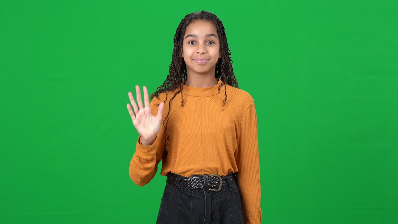 Cheerful teen African American girl waving looking at camera posing at green screen. Portrait of positive smiling beautiful teenager on chromakey background. Greeting concept.