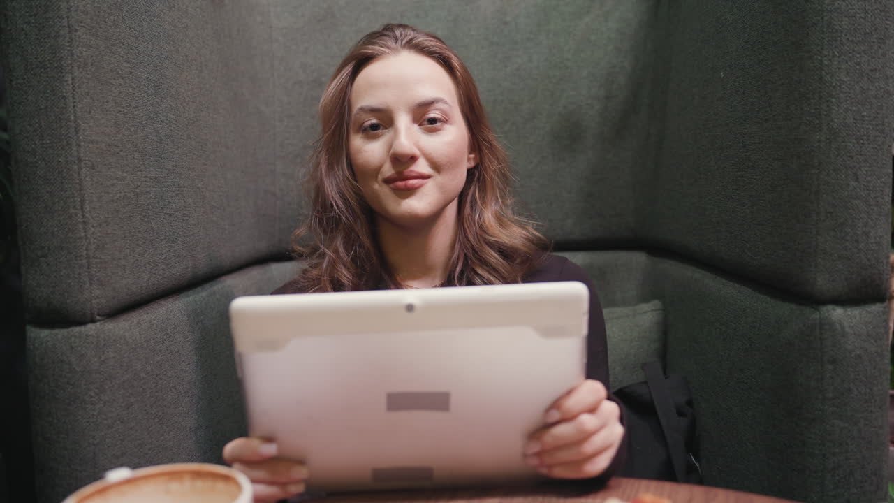 Smiling lady holds digital notepad and looks directly at camera while seated on soft cushioned booth in cozy indoor setting, exuding relaxed confidence and tech-savvy charm during calm moment