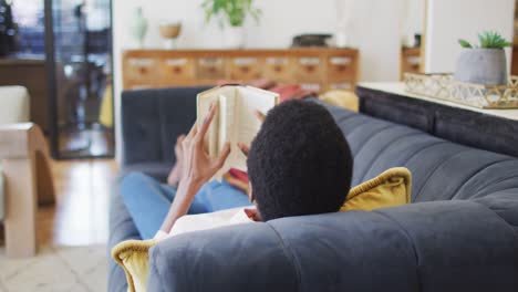 feliz mujer afroamericana acostada en el sofá en la sala de estar, leyendo un libro