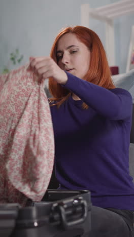 excited woman puts summer dresses into big open suitcase preparing for vacation. lady with straight red hair sits on floor in living room at home closeup