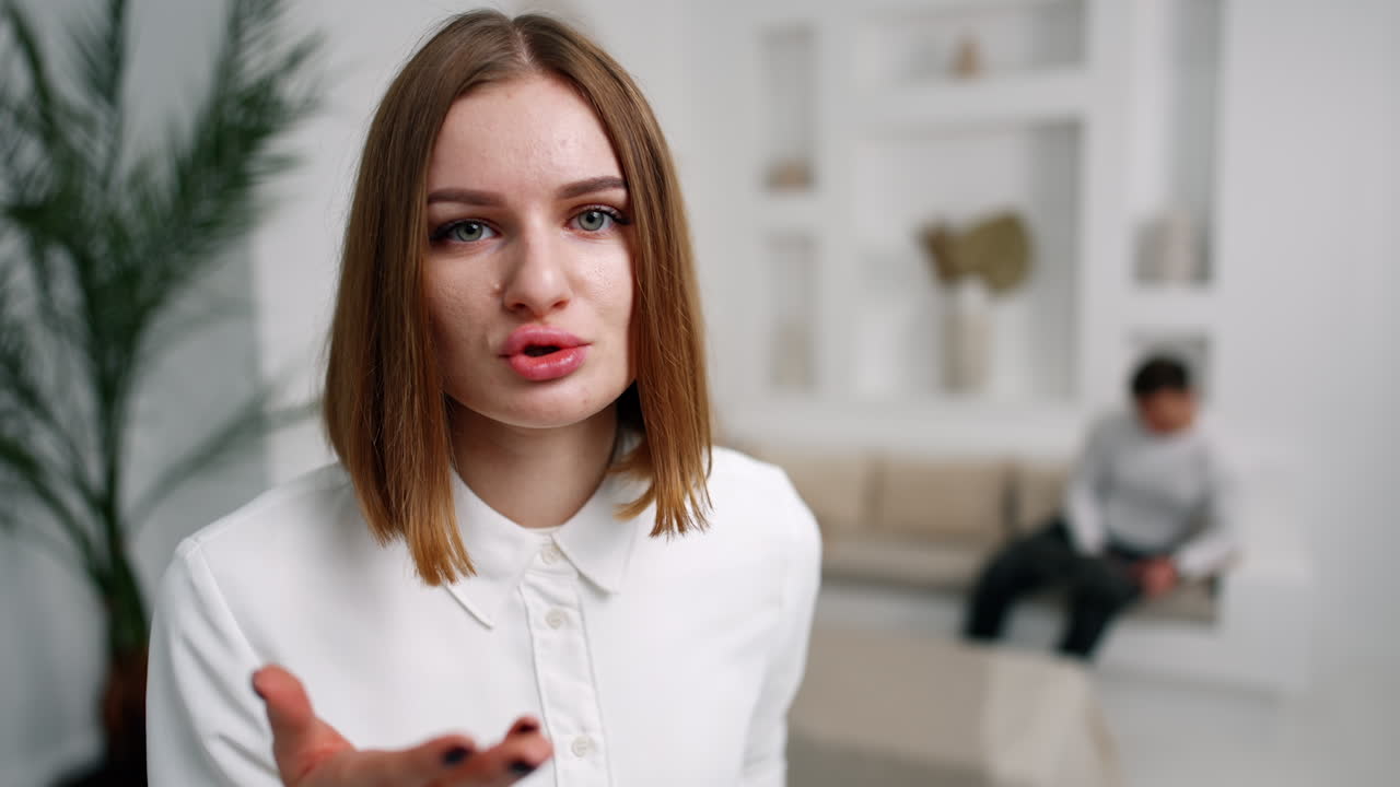 Resentful Caucasian woman looking and talking to camera. Lady is telling off to camera. Man with laptop sitting at backdrop.
