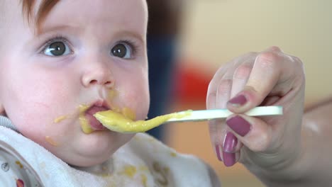 una toma de cerca de la mano de una madre empujando una cuchara llena de comida para bebés en la boca de un niño pequeño, luego el niño se pone infeliz y la escupe, toma de cerca en cámara lenta