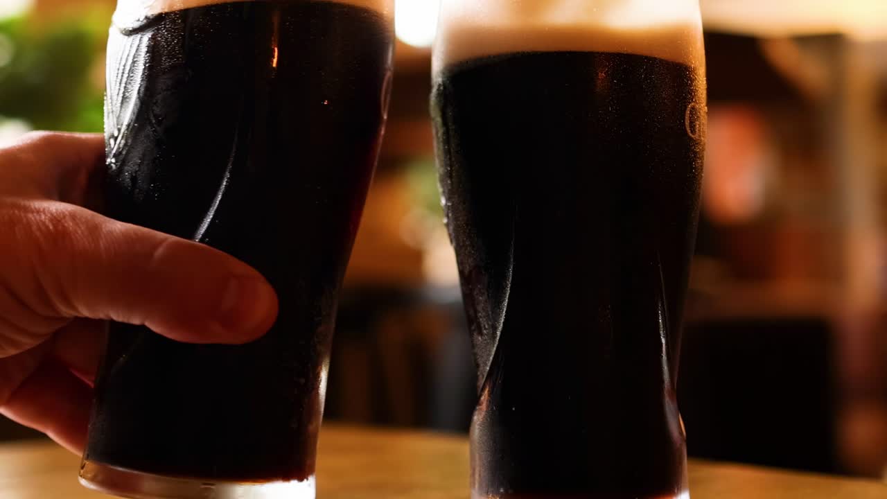 A hand raises a pint of dark beer for a toast in a warmly lit pub.