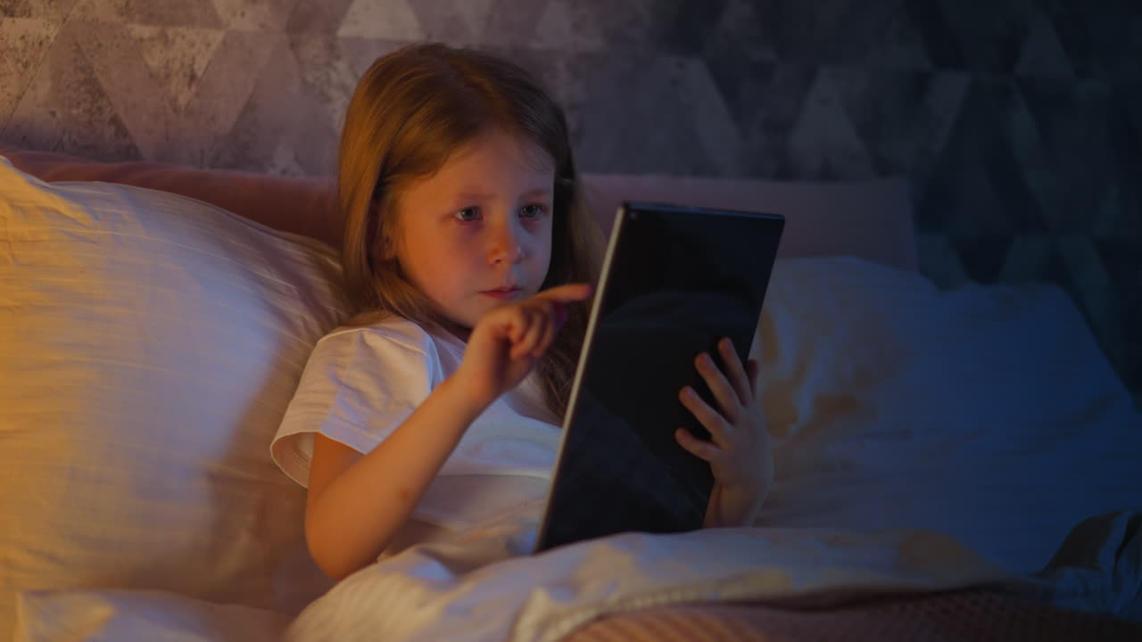 маленькая девочка использует планшет, чтобы играть в игру на кровати с подушками
