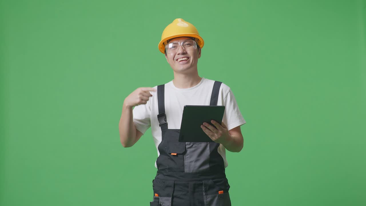 Asian Man Worker Wearing Goggles And Safety Helmet Smiling And Pointing To A Tablet In His Hand While Standing In The Green Screen Background Studio