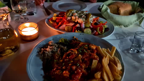 romantic, candlelight dinner at a restaurant with greek salad, seafood in sauce, and french fries - close up pullback reveal
