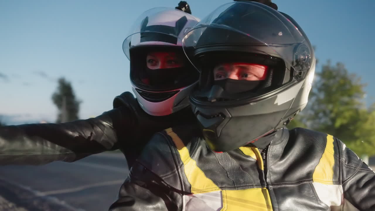 Couples ride motorcycle as lady records video with phone, gently touches husband helmet then waves, both wearing helmets and leather jackets, evening road illuminated with red light reflections