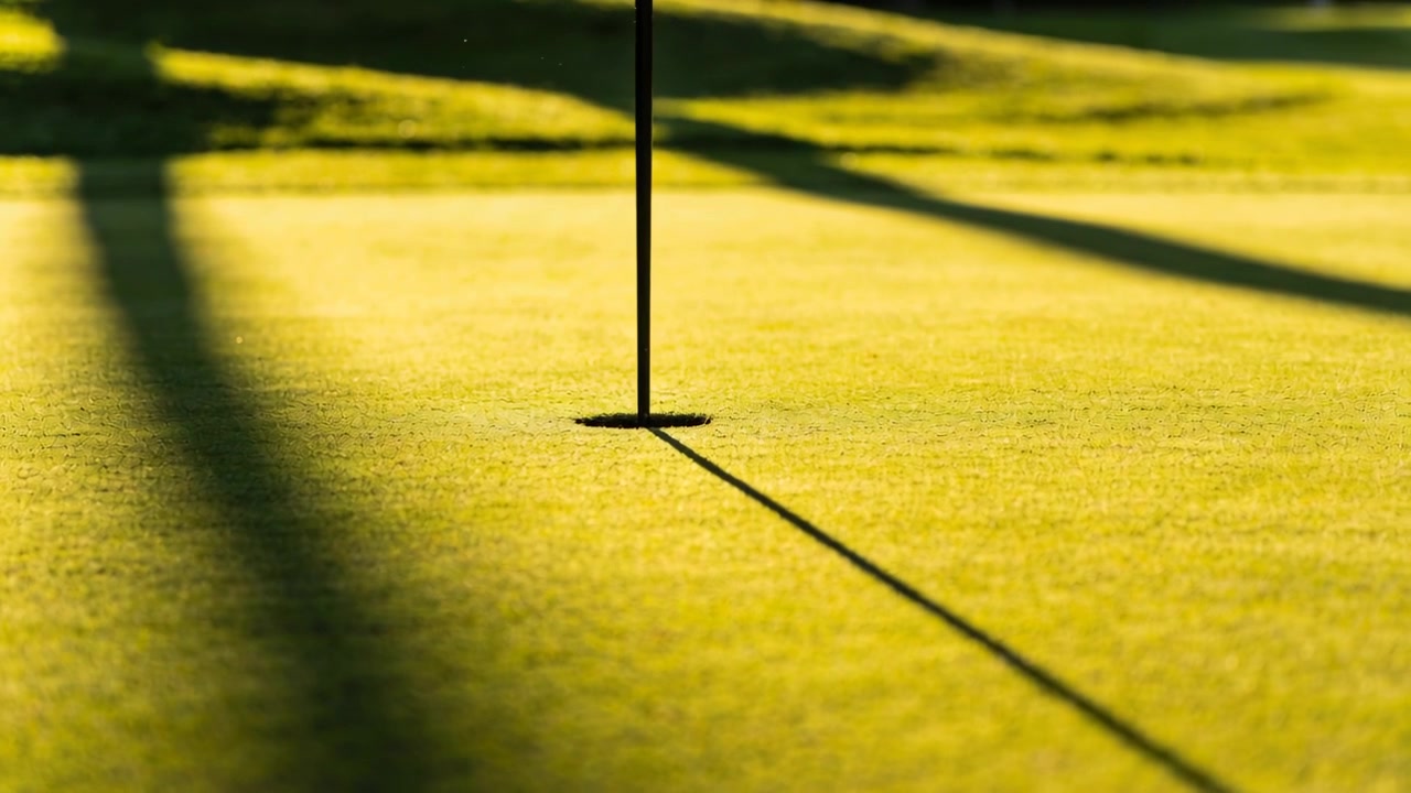 Camera kadreert verticale vlaggenstok en hole centraal op de putting green, zoomt dichterbij voor nadruk.