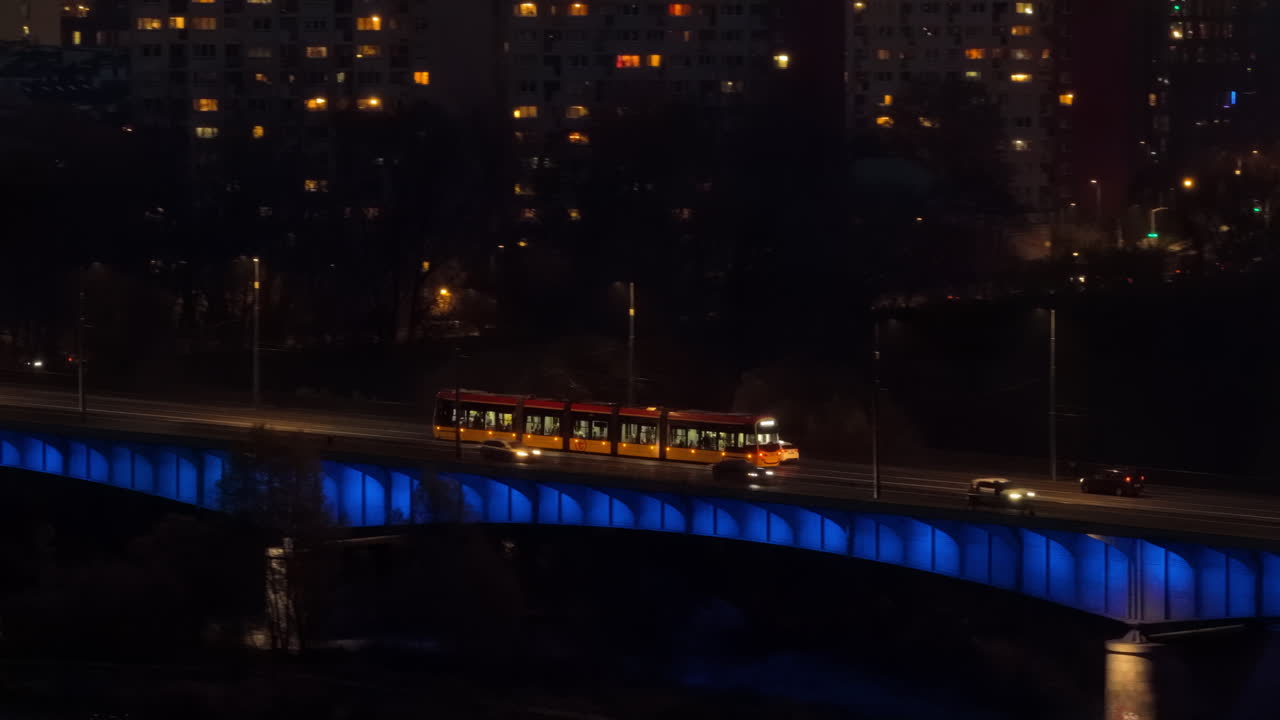 Aerial drone view of a yellow Warsaw tram traveling across the illuminated Swietokrzyski Bridge, with reflections on the Vistula River