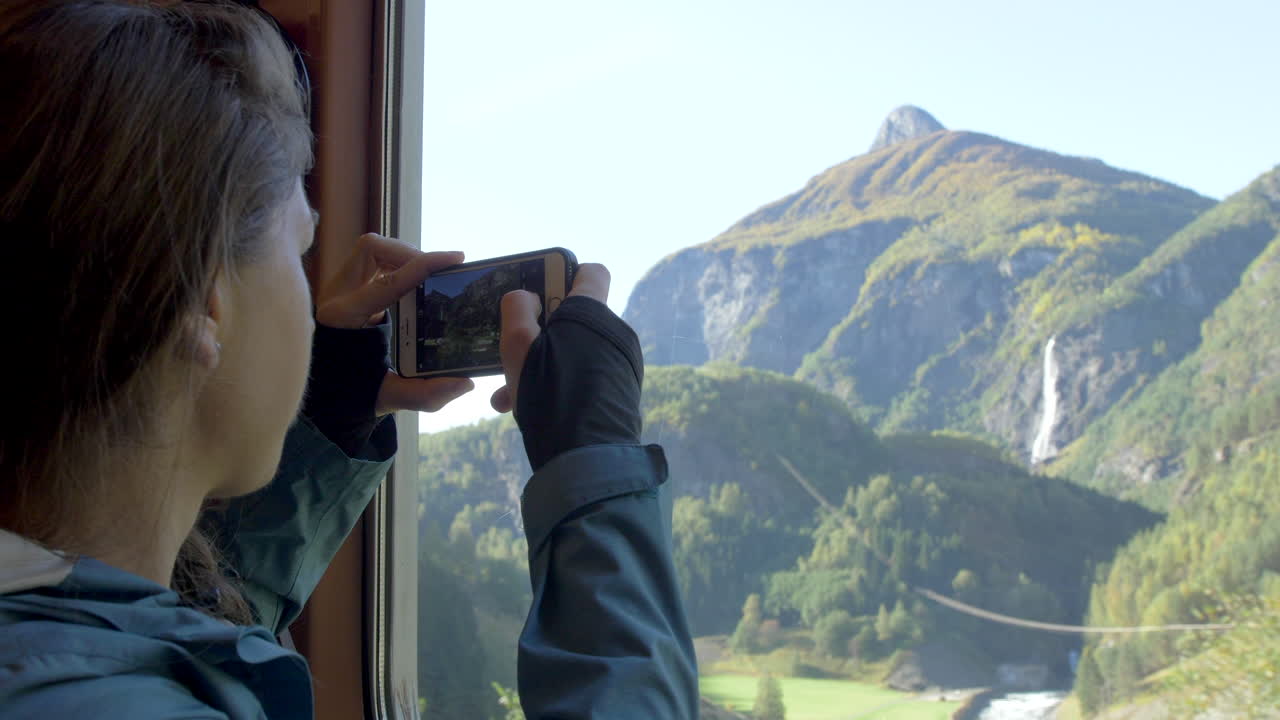 una mujer toma una foto con su teléfono de un pintoresco paisaje montañoso fuera de la ventana de un tren en noruega