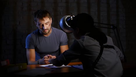 closeup criminal in handcuffs is required to write confessionary statements man dressed in a gray tshirt sitting at the table it stacked folders and evidence woman detective shows a finger where and what to write the offender submits close shot