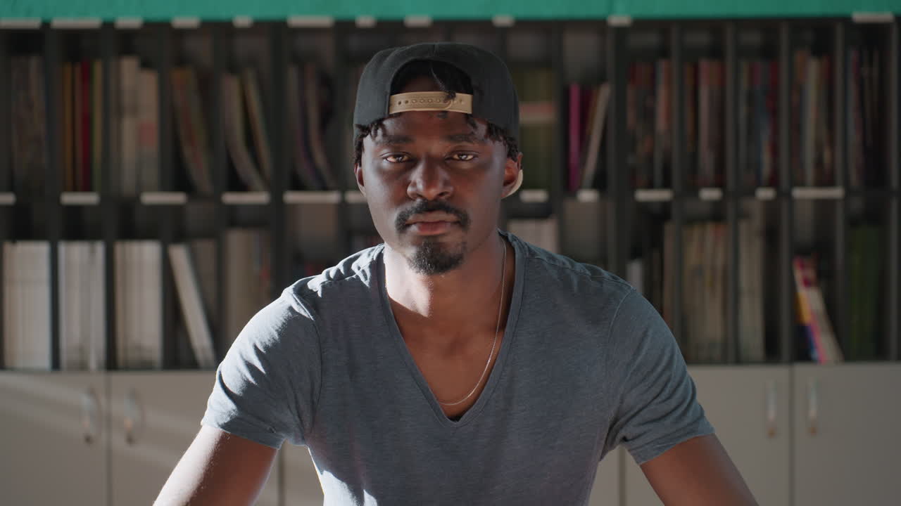 Boy wearing cap sits at library table leaning forward with stack of books, slowly lifting head and eyes closed after reading, teal wall and shelves behind, warm light creating contemplative mood