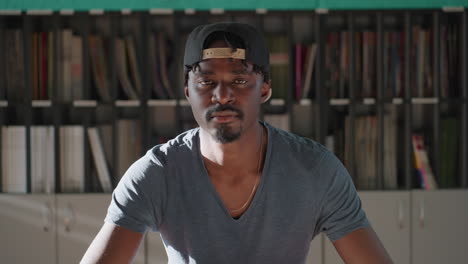 boy wearing cap sits at library table leaning forward with stack of books, slowly lifting head and eyes closed after reading, teal wall and shelves behind, warm light creating contemplative mood