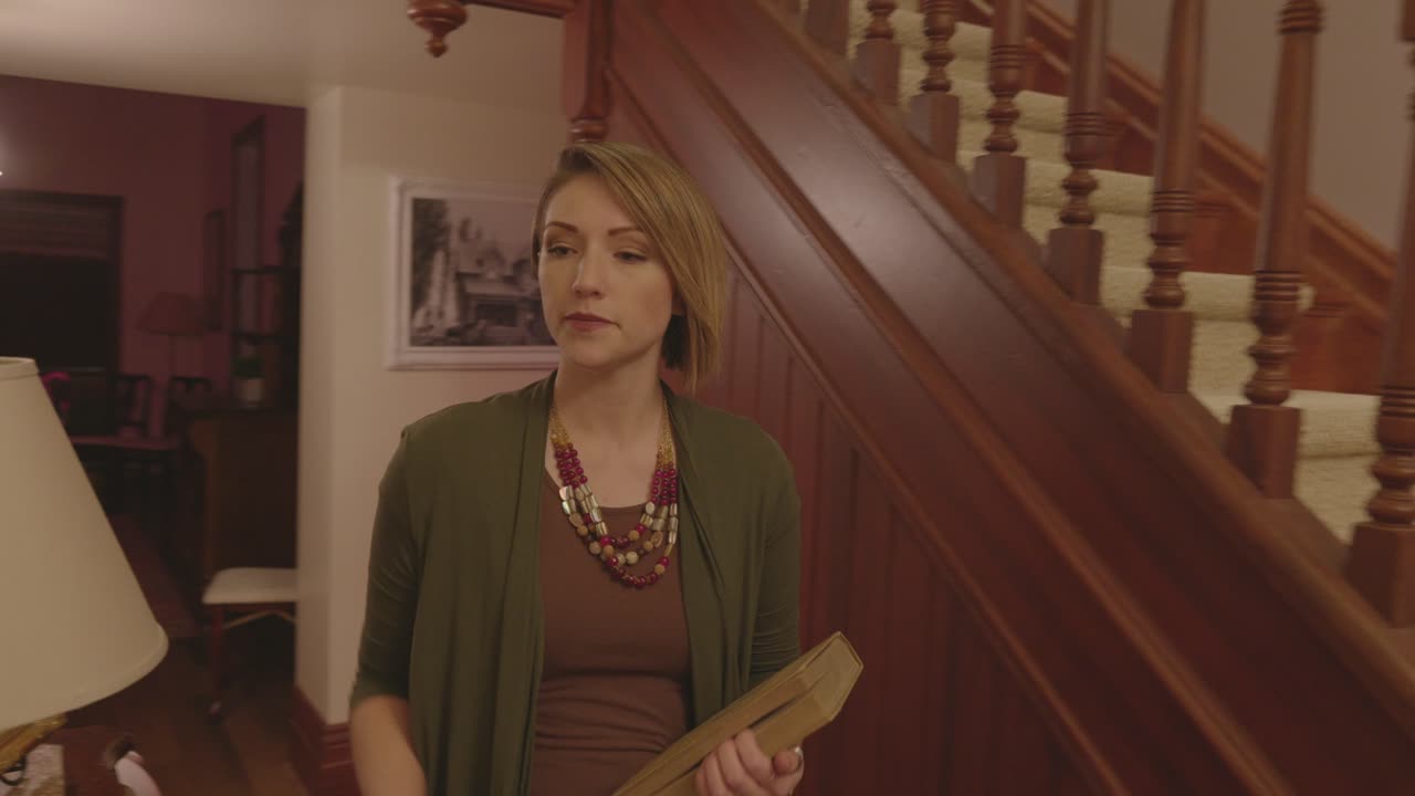 A woman is walking down a hallway and up the stairs holding a book. She turns off the lights on the way up the stairs