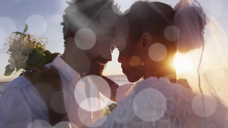 animation of light spots over happy african american bride and groom embracing on beach at wedding