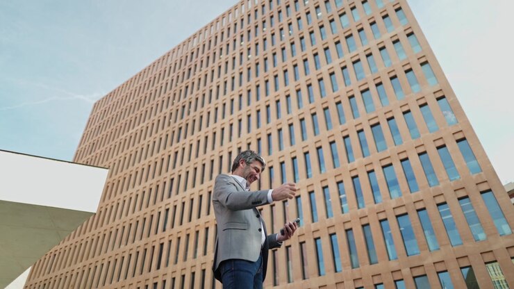 Uomo d'affari frustrato riceve cattive notizie sullo smartphone fuori da un ufficio moderno.