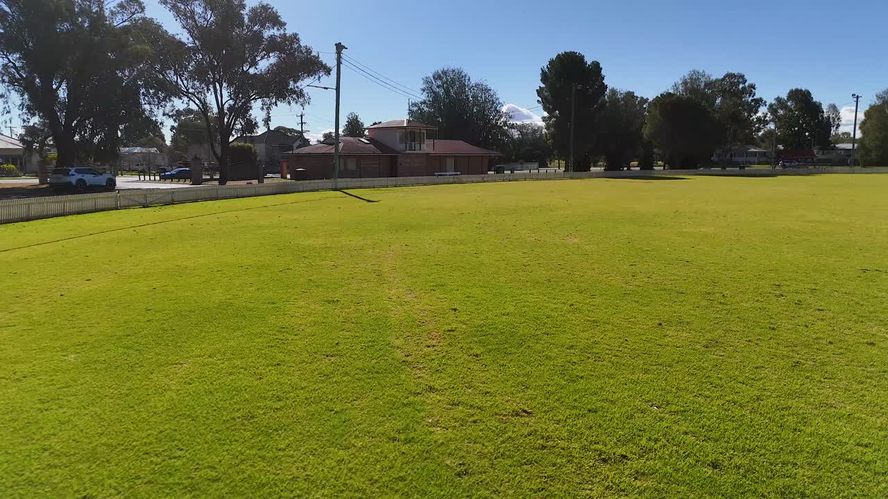 A steady camera moves forward across a bright green sports ground under clear blue skies, with suburban houses and trees in the background