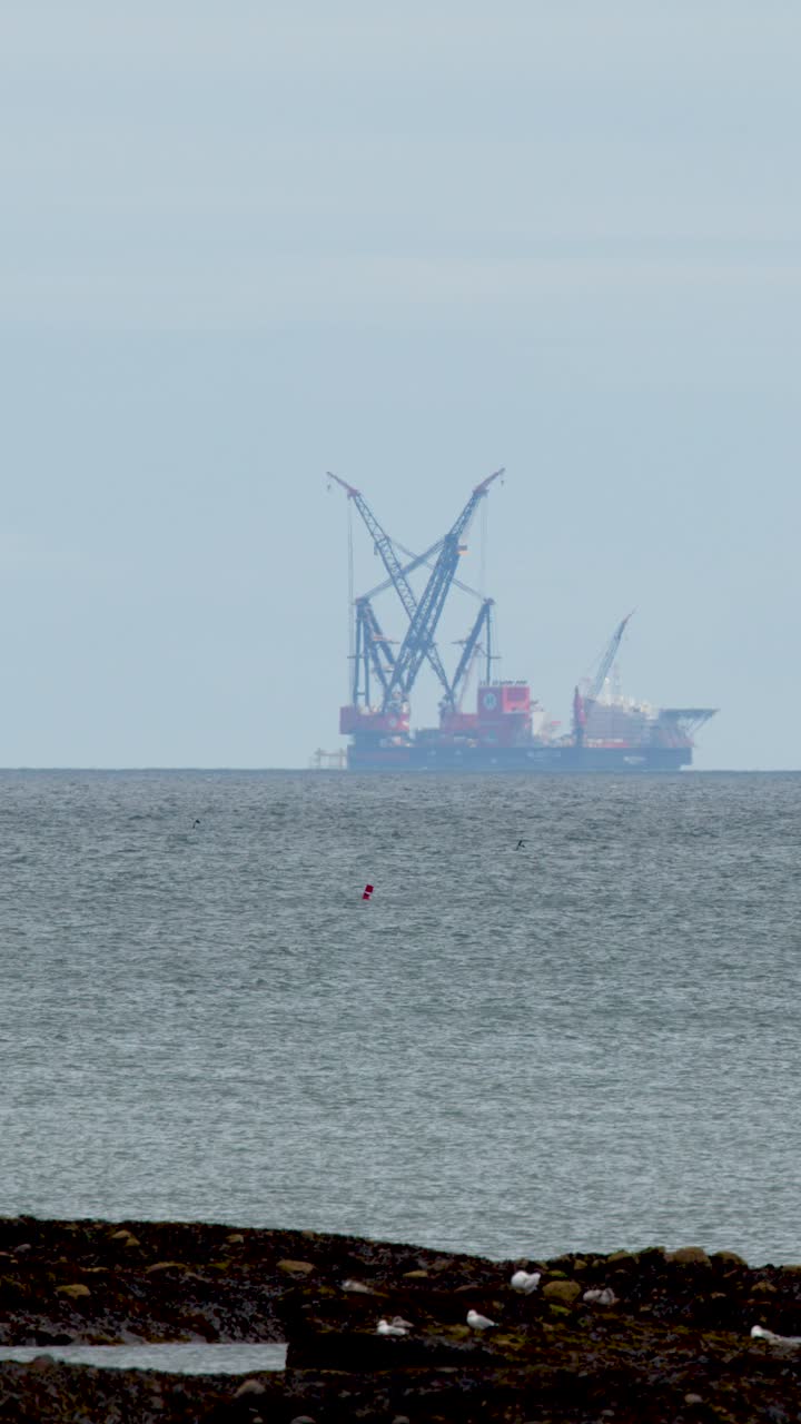 Large offshore industrial barge with cranes travels across calm sea under overcast daylight, distant perspective