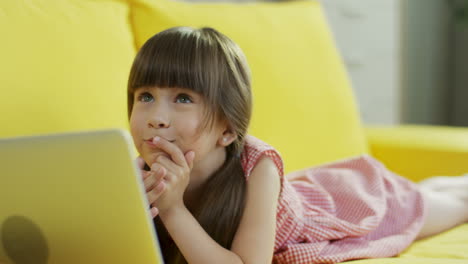 Niñita-Linda-Jugando-Con-Una-Laptop-Mientras-Está-Acostada-En-Un-Sofá-Amarillo-En-La-Sala-De-Estar