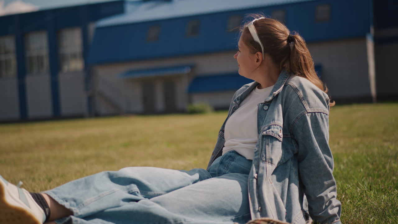 Donna in jeans seduta pacificamente. Donna serena in abbigliamento casual che si gode l'ambiente soleggiato del campus. Persona vestita casualmente in jeans seduta sull'erba vicino a un edificio universitario sotto la calda luce del sole.