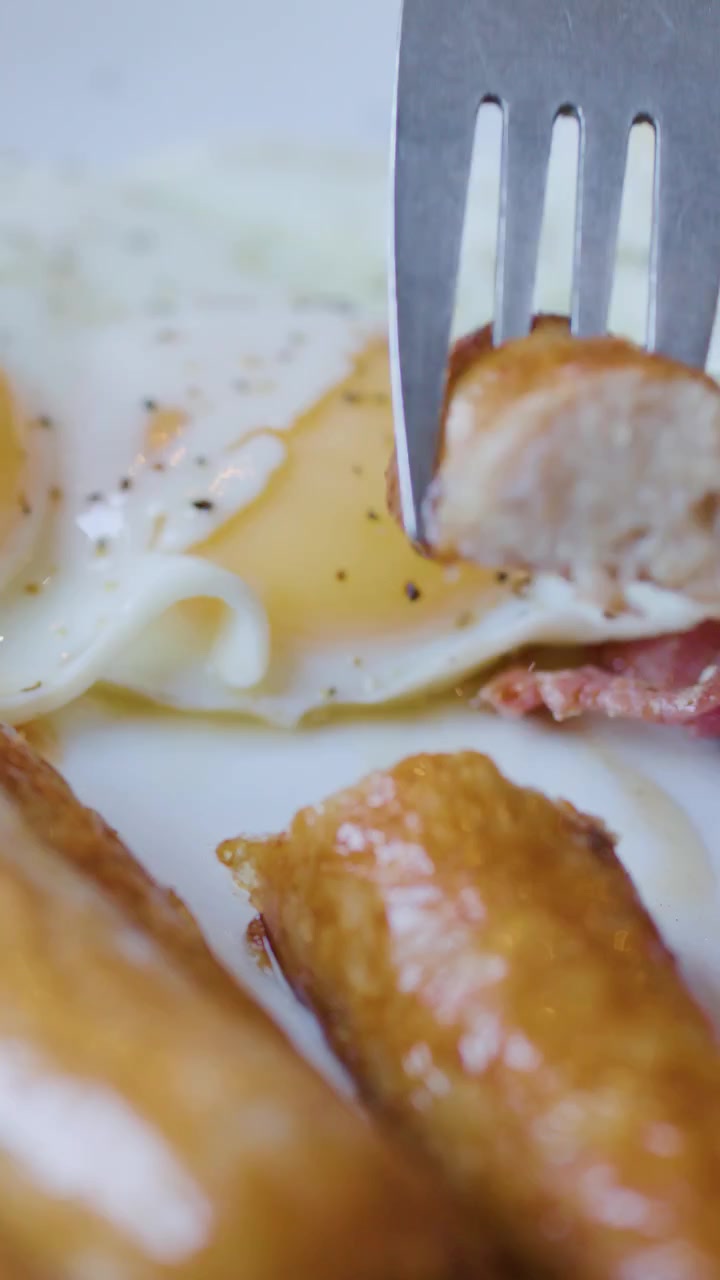 Una mano affetta salsiccia con coltello e forchetta, servita con uova e pancetta sotto luce naturale