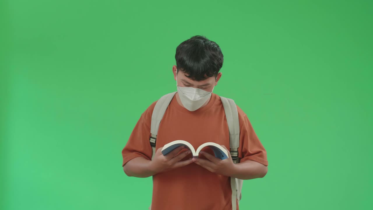 The Front View Of Asian Boy Student Wearing A Mask, Backpack And Brown T-Shirt Reading A Book While Walking To School On Green Screen Chroma Key.