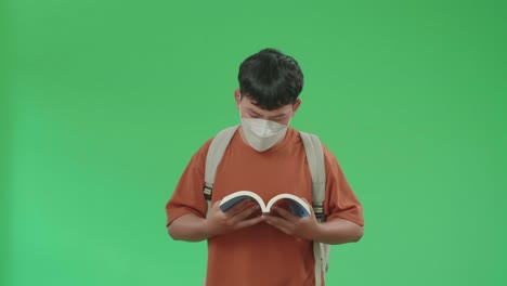 the front view of asian boy student wearing a mask, backpack and brown t-shirt reading a book while walking to school on green screen chroma key.