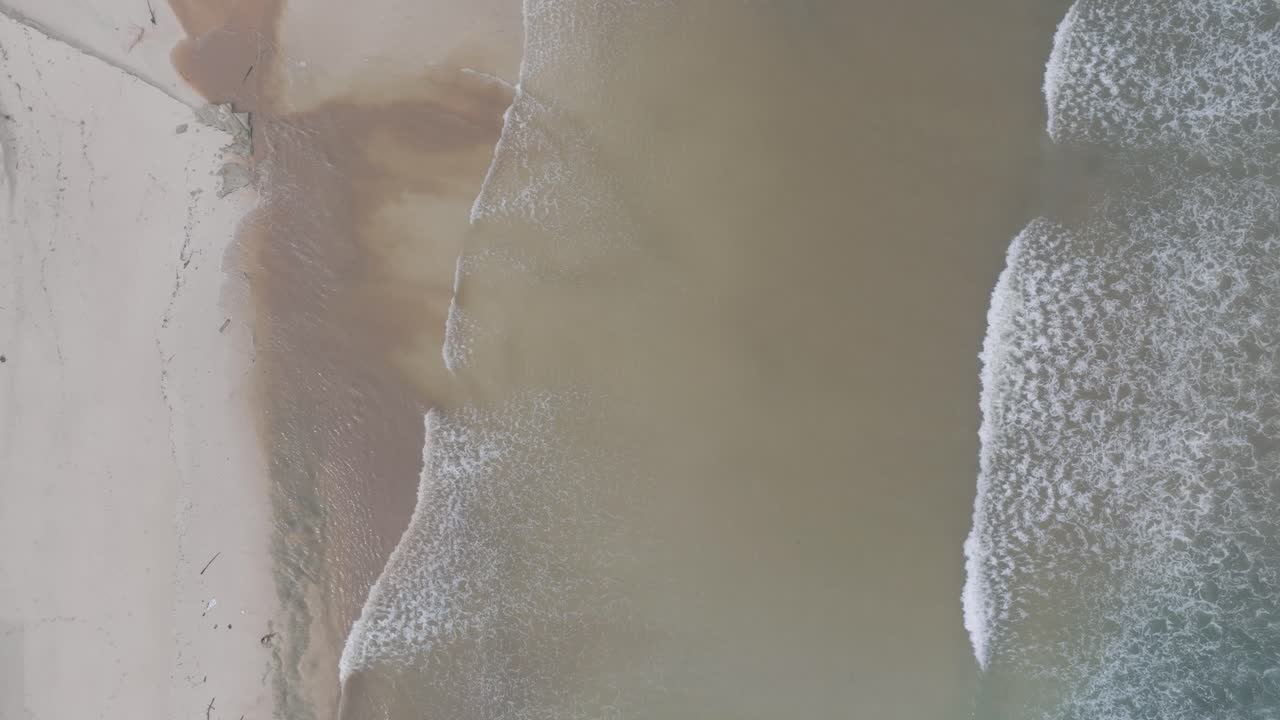 drone de arriba hacia abajo disparó pequeñas olas de costa que se rompen en la arena y se mezclan con la escorrentía de aguas residuales de ríos fangosos en bali, indonesia