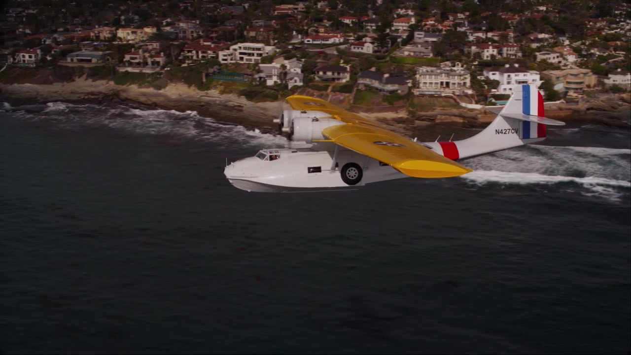 Spektakulært luft-til-luft dronebilde av et historisk PBY Catalina sjøfly fra andre verdenskrig som cruiser over San Diegos kystlinje, med det ikoniske Hotel del Coronado og byens skyline synlig i bakgrunnen
