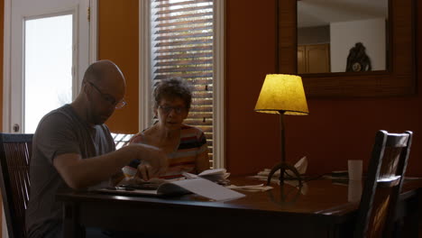 a middle-aged man and his senior mother sit together at a dining table in a warmly lit home, sorting through papers and bills. son counts receipts, while mother is onlooking. 24 fps.