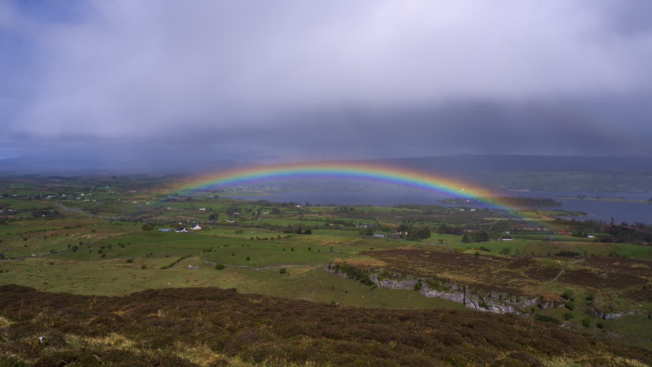 Timelapse af et landligt dalområde med en passerende regnstorm og fremkomsten af en regnbue, bakker og en sø i det fjerne på en overskyet og solrig forårsdag set fra Carrowkeel i County Sligo i Irland