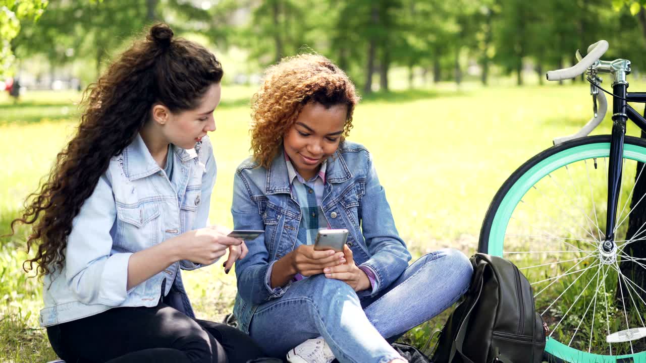 Young African American woman is shopping online making payment with smartphone while her friend in holding credit card, girls are sitting on lawn in park in summer.
