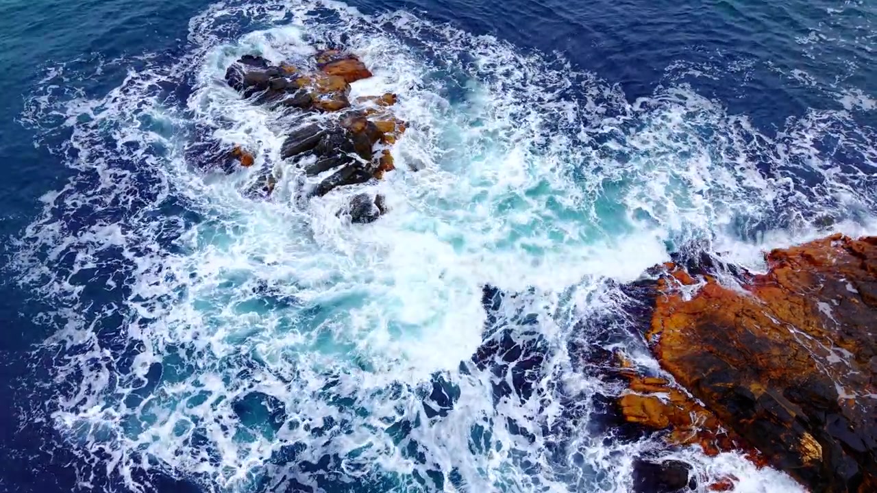 Close-up vanuit de lucht van schuimende golf die breekt bij rots. Bovenaanzicht van drone vangt diepblauw water, wit schuim en donkerrode rotsblok