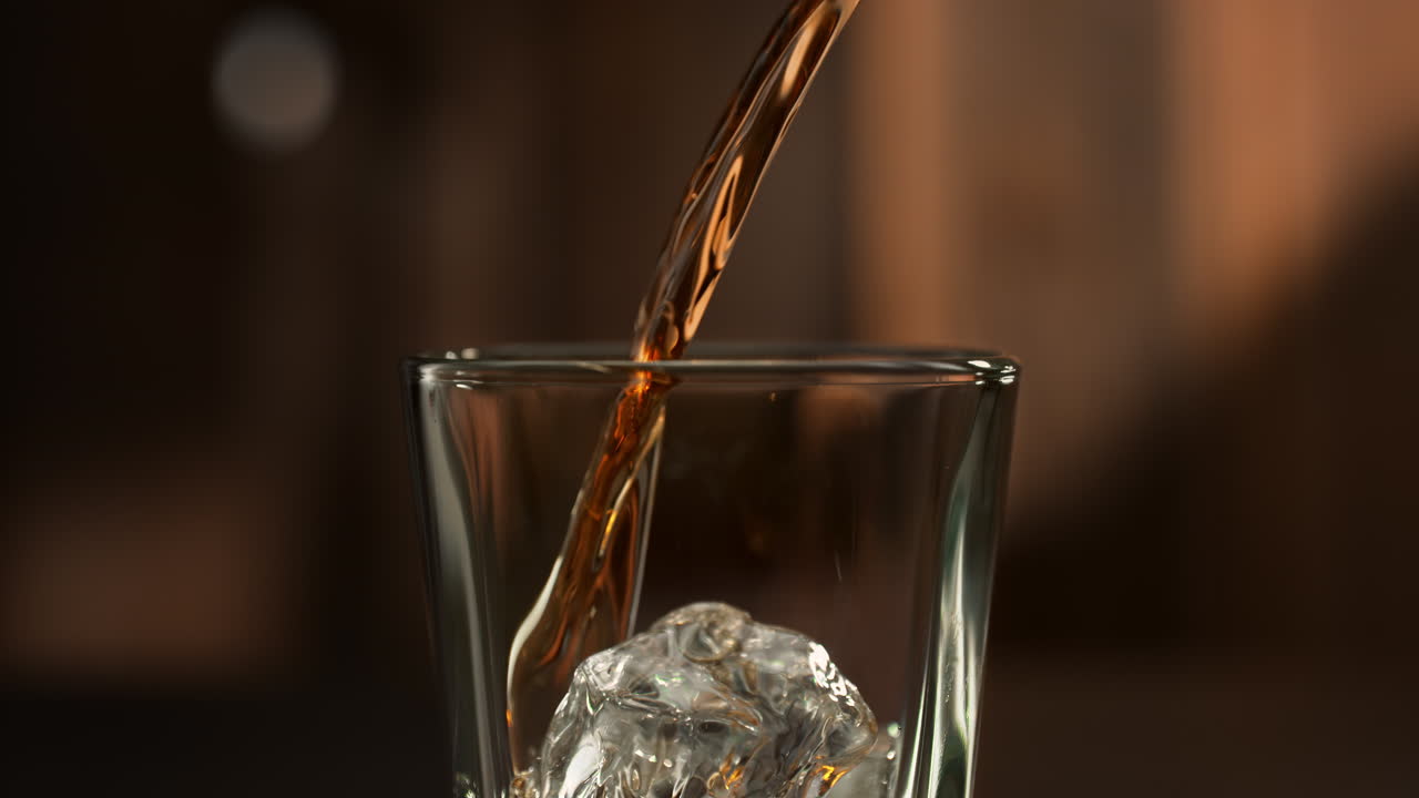una corriente de whisky fluye hacia el vaso salpicando sobre el cubo de hielo en cámara lenta