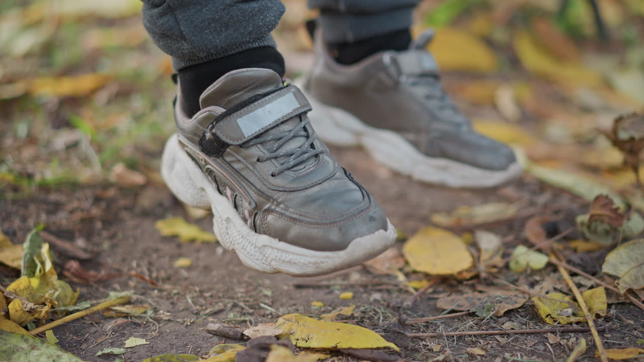 child legs swinging slowly above fallen autumn leaves scattered on dirt path with gentle drift of foliage beneath chunky sneakers evoking tranquil seasonal outdoor moment with soft bokeh