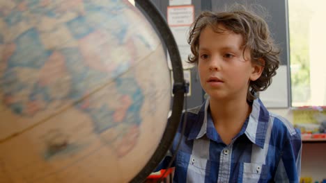 vista frontal de un escolar caucásico estudiando el globo en el escritorio en el aula de la escuela 4k