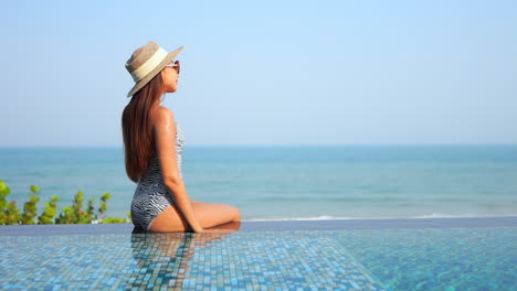 Young-asian-woman-in-swimsuit-sitting-on-infinity-pool-border-looking-around-and-enjoying-on-hot-summer-day,-slow-motion