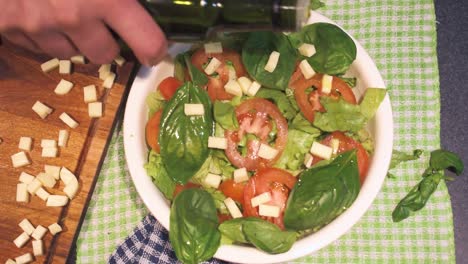 manos haciendo lechuga y tomate con albahaca, añadiendo aceite de oliva en un recipiente blanco.