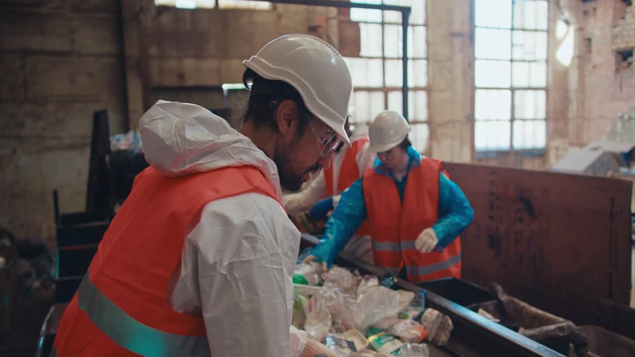 un hombre moreno con un uniforme especial y un chaleco naranja recicla la basura y selecciona botellas de plástico de un color específico junto con sus empleados cerca de una cinta transportadora en una planta de reciclaje de residuos