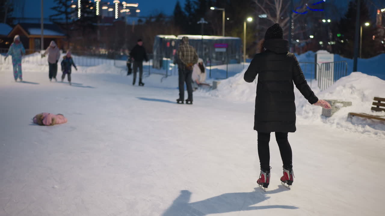 Skater gliding gracefully on outdoor ice rink at night wearing black coat, red skates, patterned mittens, surrounded by other skaters enjoying winter activity under glowing lights and snowy festive park